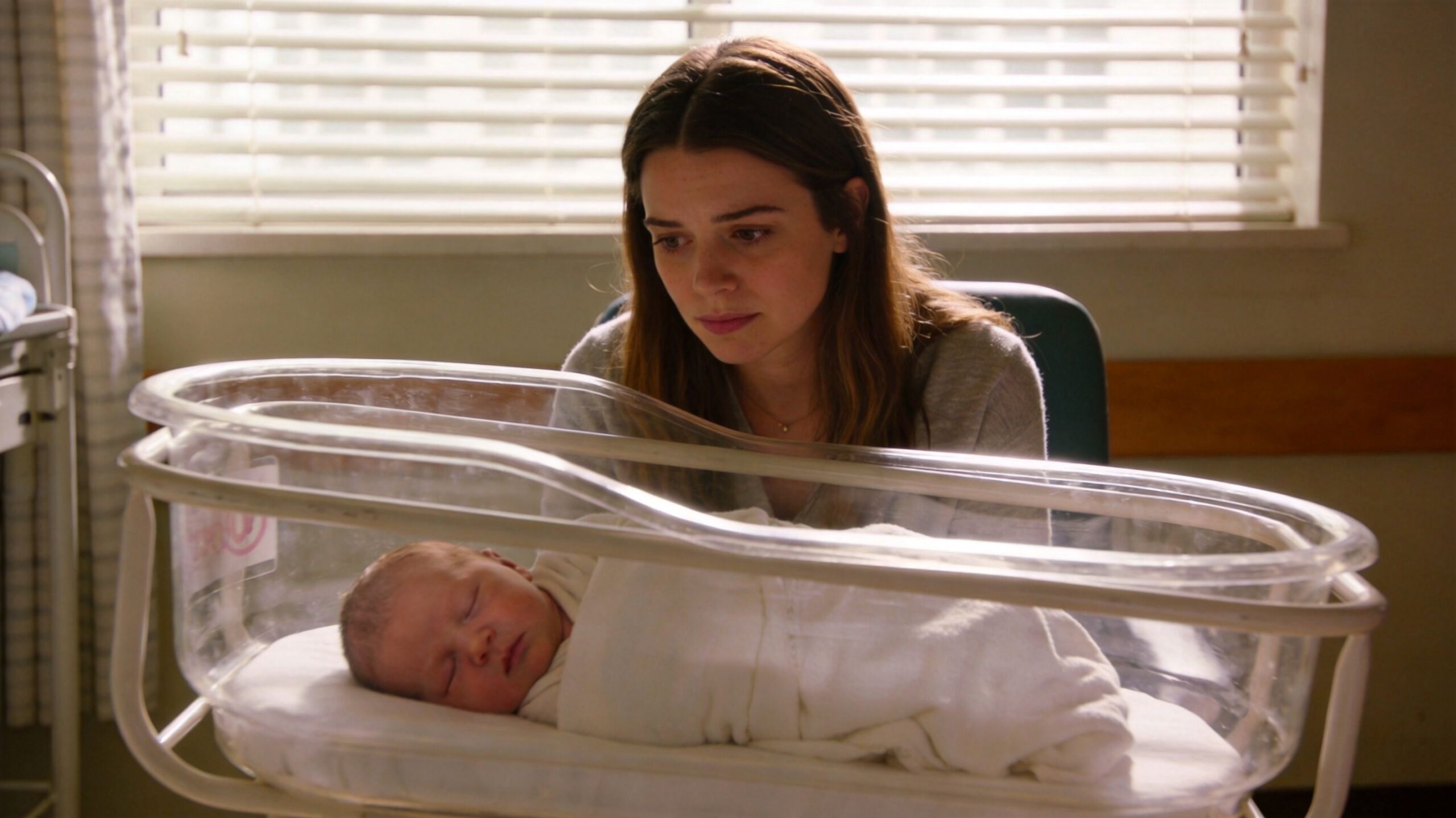 A concerned mother watches over her newborn baby sleeping peacefully in a clear hospital bassinet.