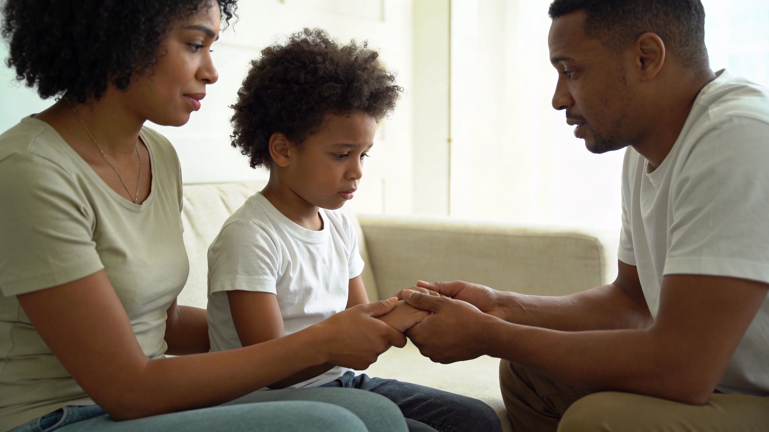 A concerned Black mother and father comforting their young son while holding his hands on a sofa.